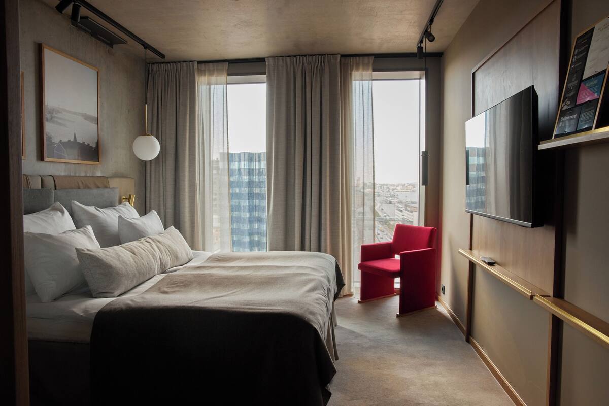 Habitación de hotel con una cama grande, una silla roja, un televisor y vistas a la ciudad.