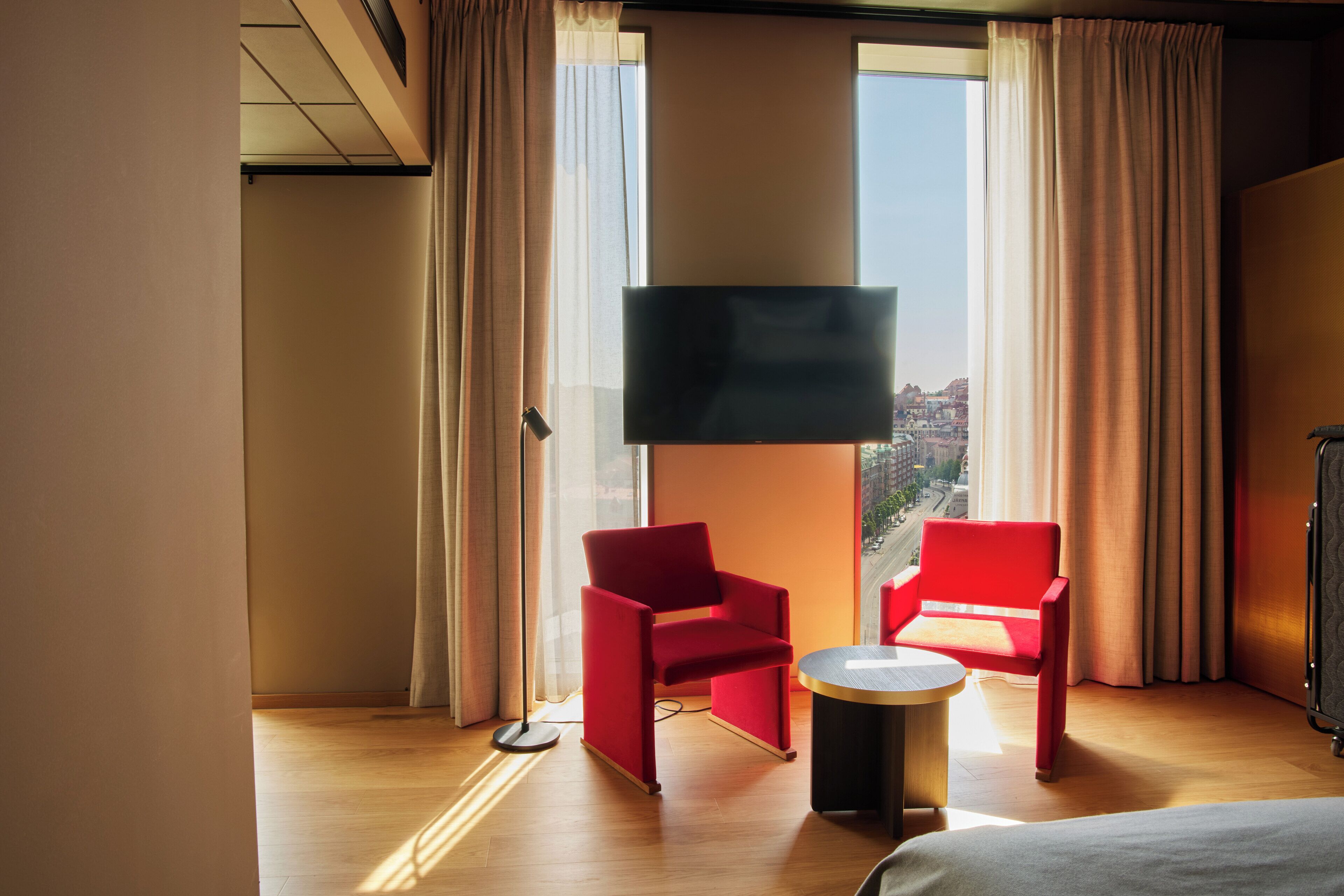 A hotel room with a large window, two red chairs, a small round table, and a flat-screen TV mounted on the wall.
