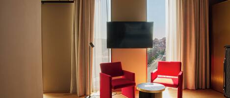 A hotel room with a large window, two red chairs, a small round table, and a flat-screen TV mounted on the wall.