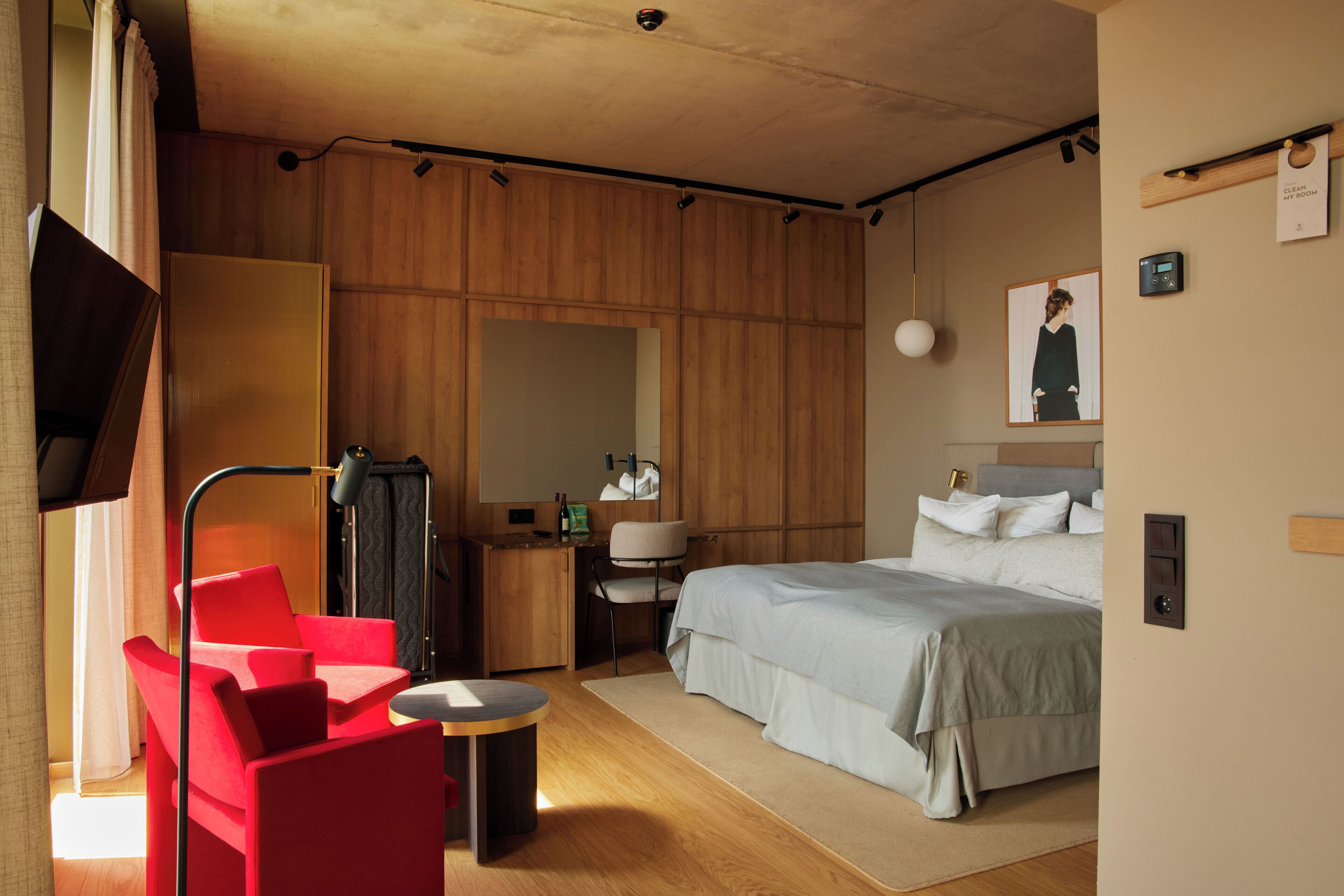 A modern hotel room with a wooden wardrobe, a bed with white bedding, a red armchair, a small round table, and a mirror on the wall.