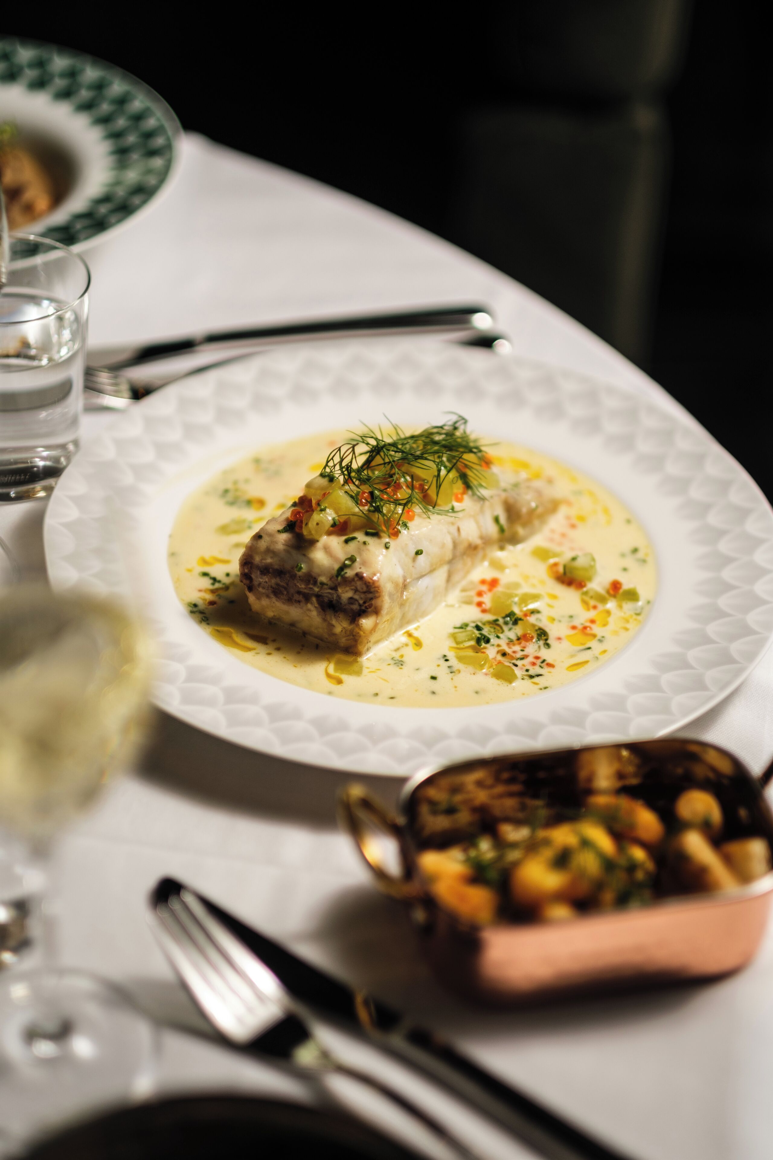 Un plato blanco con un guiso de pescado, un vaso de agua y un cuenco pequeño de verduras sobre una mesa.