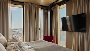 Habitación de hotel con cama, una silla roja, televisión y vista a un paisaje urbano a través de amplias ventanas.