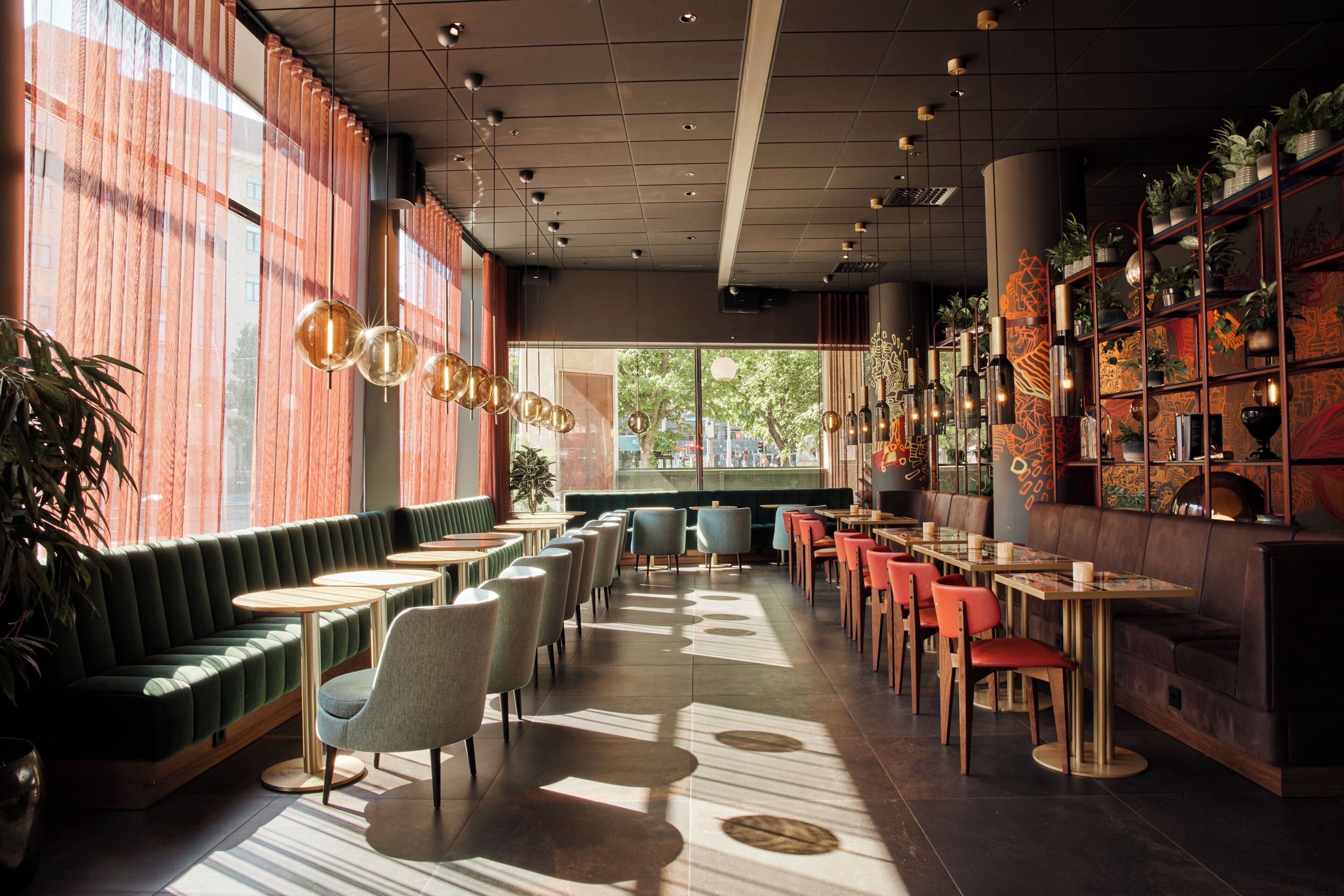 Un interior de restaurante moderno con una zona de asientos en cabinas verdes, mesas redondas y una pared decorada con plantas y obras de arte.