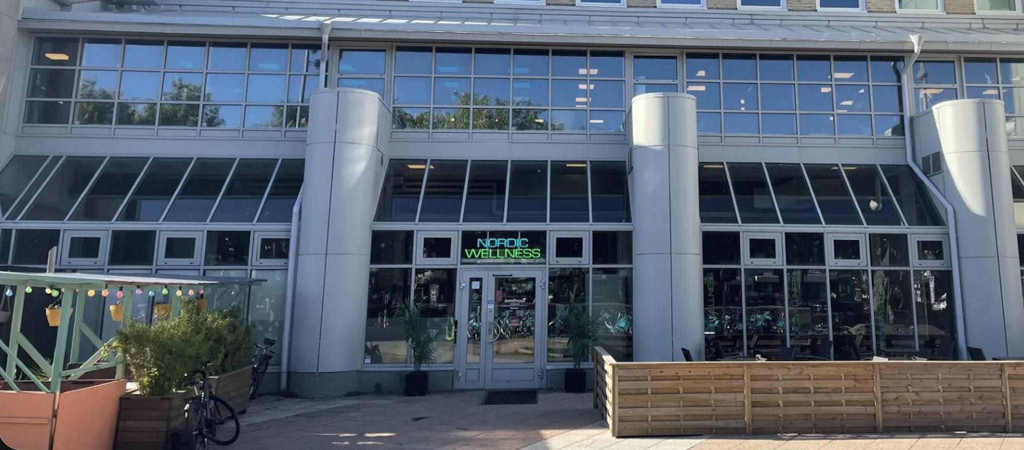 A modern building with a glass facade, a sign reading 'Nordic Wellness,' and a wooden bench outside.
