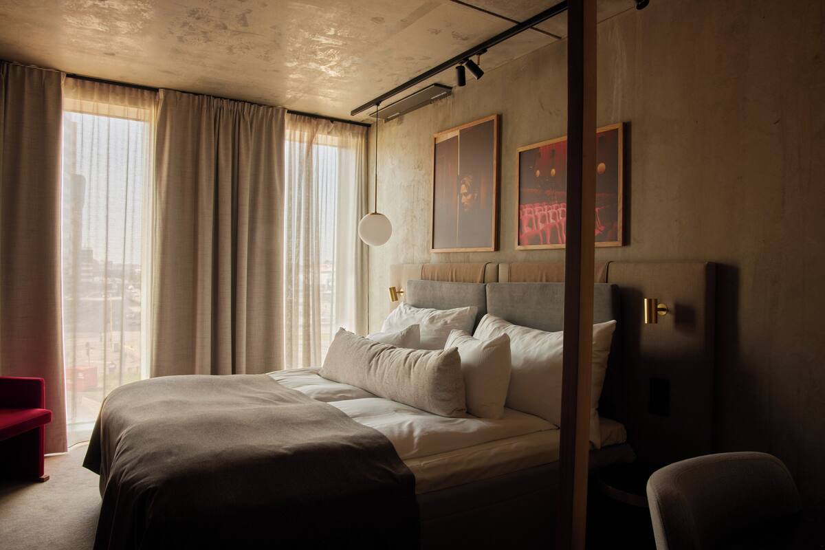 Habitación de hotel moderna con una cama grande, una silla roja y vistas a la ciudad a través de la ventana.