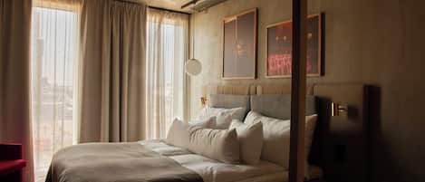 A modern hotel room with a large bed, a red chair, and a city view through the window.