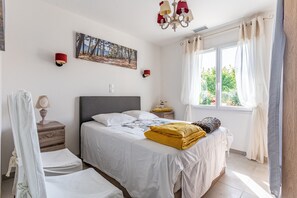 A bedroom with a bed, bedside tables, a nightstand, a window with curtains, and a chandelier.
