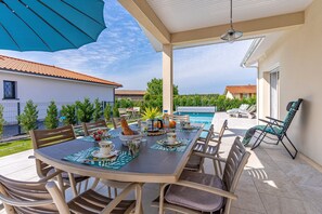 An outdoor patio with a dining table set for four, surrounded by chairs, a pool, and a covered area with a ceiling fan.