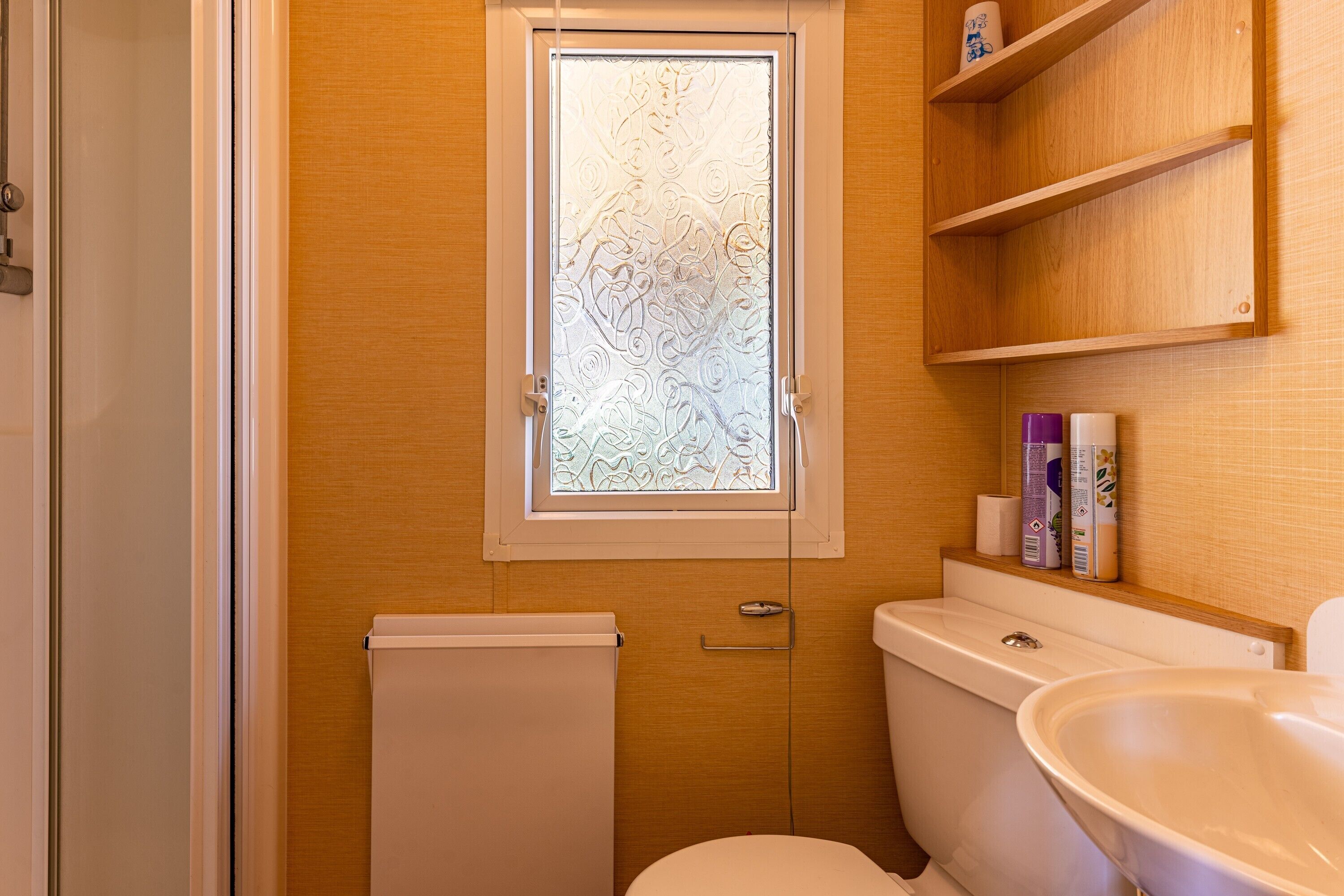 A bathroom with a toilet, a sink, a window with frosted glass, and a shelf with toiletries.