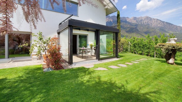 Une maison moderne dotée d’une véranda en verre, d’une pelouse bien entretenue et offrant une vue sur les montagnes.