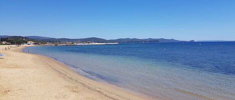 Ein Strand mit klarem Wasser, einem Sandufer und hügeliger Landschaft in der Ferne.