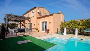 Et hus med swimmingpool, terrasse med stole og pergola.