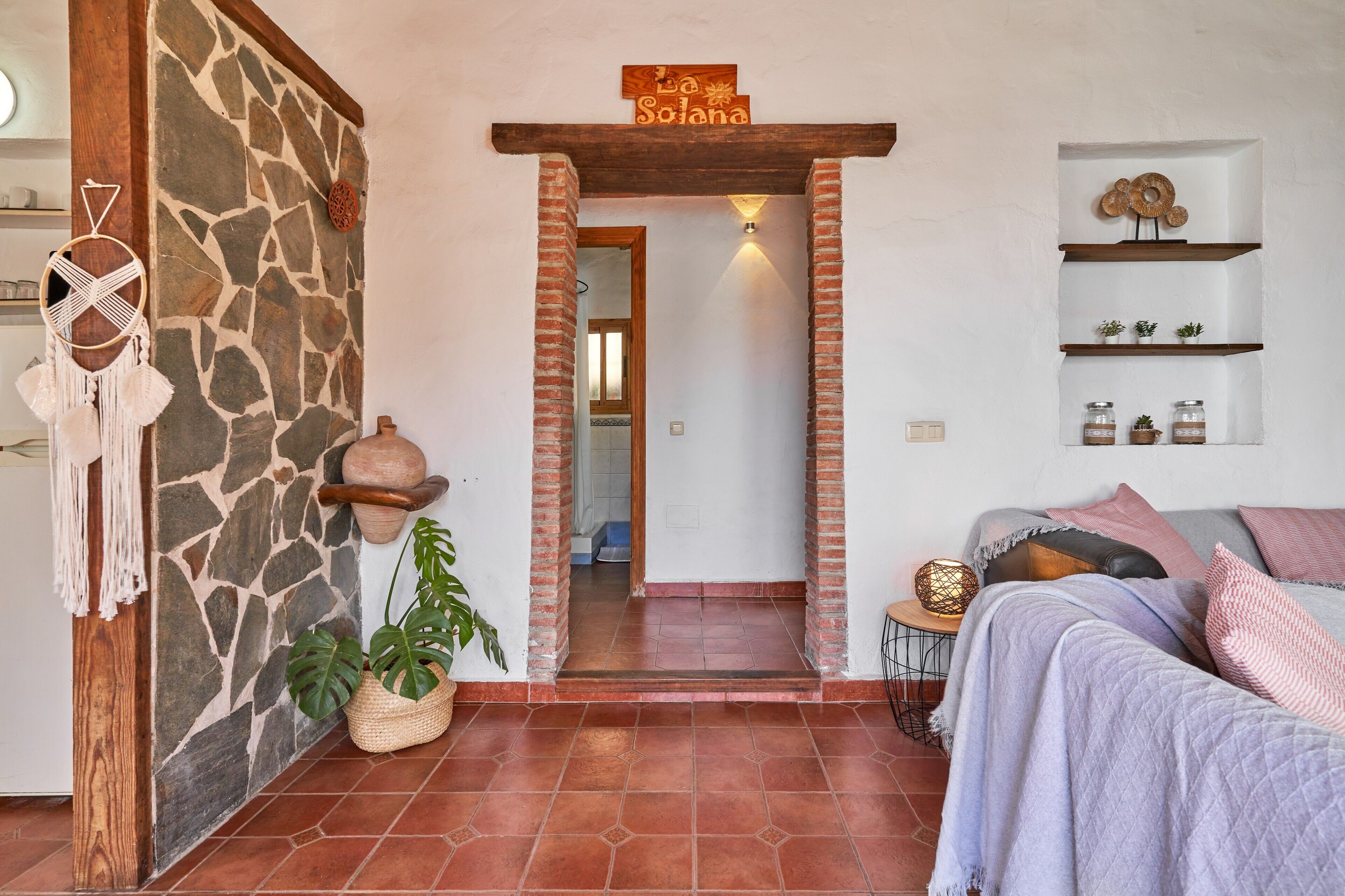 Un salón acogedor con una pared de piedra, una puerta de madera con la inscripción 'La Solana', un sofá con una manta y un estante con objetos decorativos.