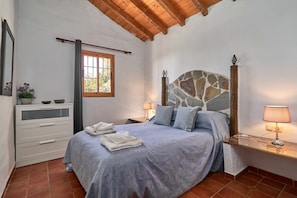 Un dormitorio con cabecera de piedra, una cama con cubrecama gris, una mesita de noche con lámpara, un aparador con espejo y una ventana con cortinas.