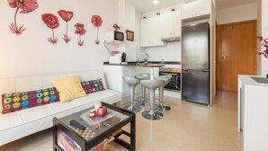 A modern living room with a sofa, coffee table, and bar stools. There is a kitchen area with a refrigerator, microwave, and cabinets.
