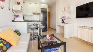 A modern living room with a sofa, a coffee table, a TV, and a kitchen area with white cabinets and appliances.