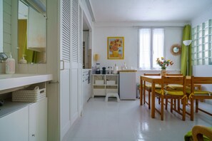 Une cuisine avec des meubles blancs, une table à manger en bois avec des chaises, un petit réfrigérateur et un miroir.