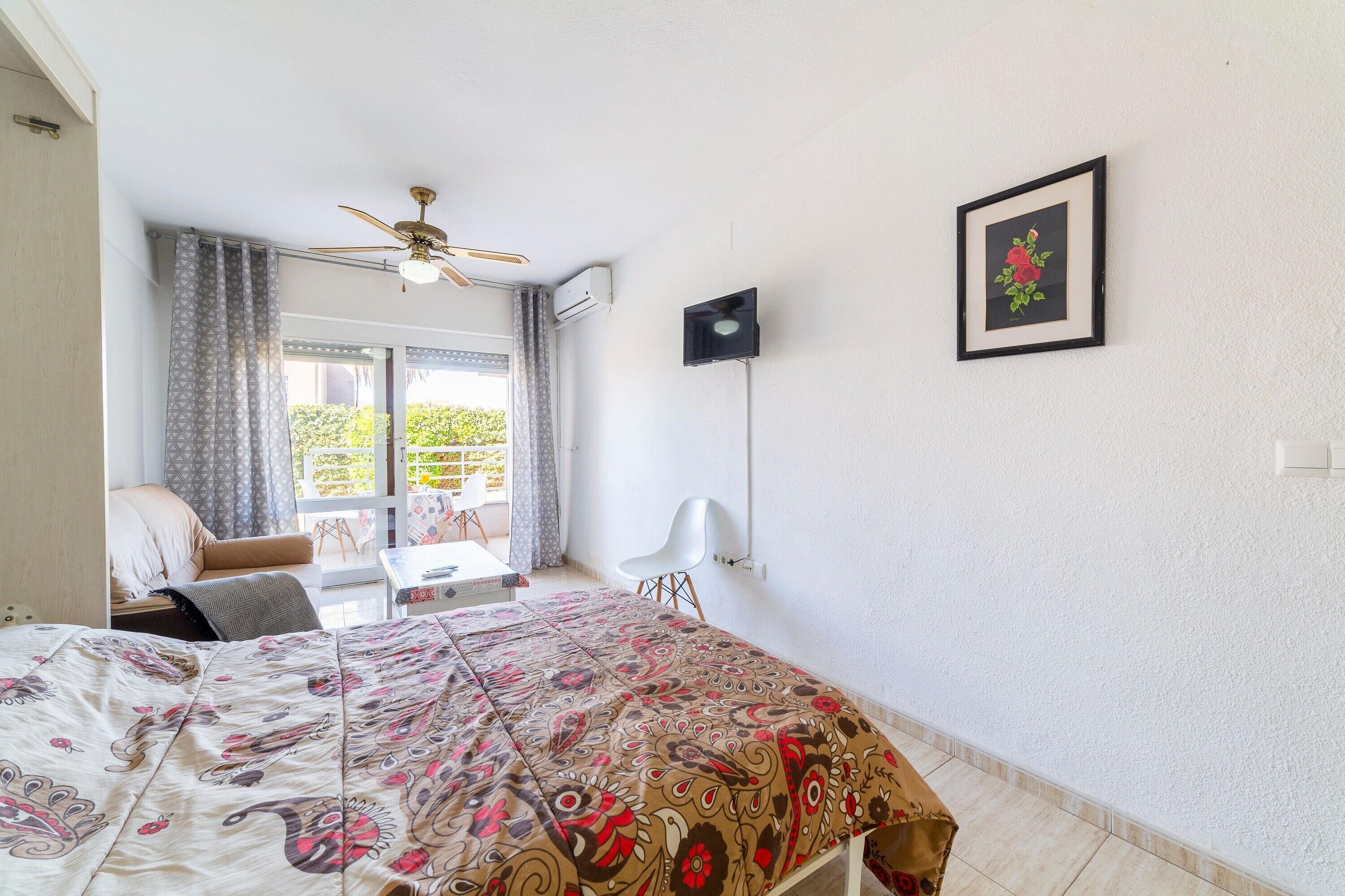 Une chambre à coucher avec un lit, un ventilateur de plafond, une fenêtre avec des rideaux, une chaise et un tableau encadré au mur.