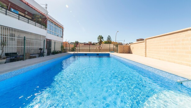 A swimming pool with a clear blue water surface, surrounded by a white building with a red roof and a brick wall. - Studio 'Gaviotas 4' with Communal Pool, Wi-Fi, and Air Conditioning (Aguadulce)