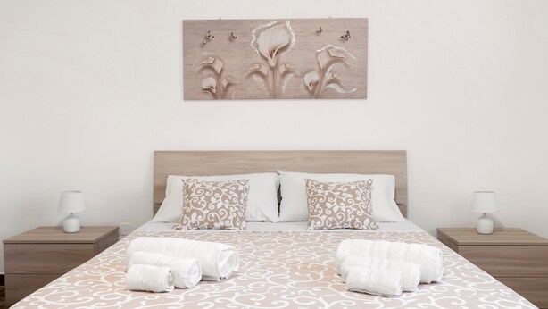Una cama bien tendida con ropa de cama estampada, dos mesitas de noche con lámparas, y una obra de arte colgada en la pared sobre la cama.