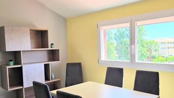 Een eetruimte met een witte tafel en vier stoelen, een houten schapenkast, en een raam met uitzicht op groen en gebouwen.