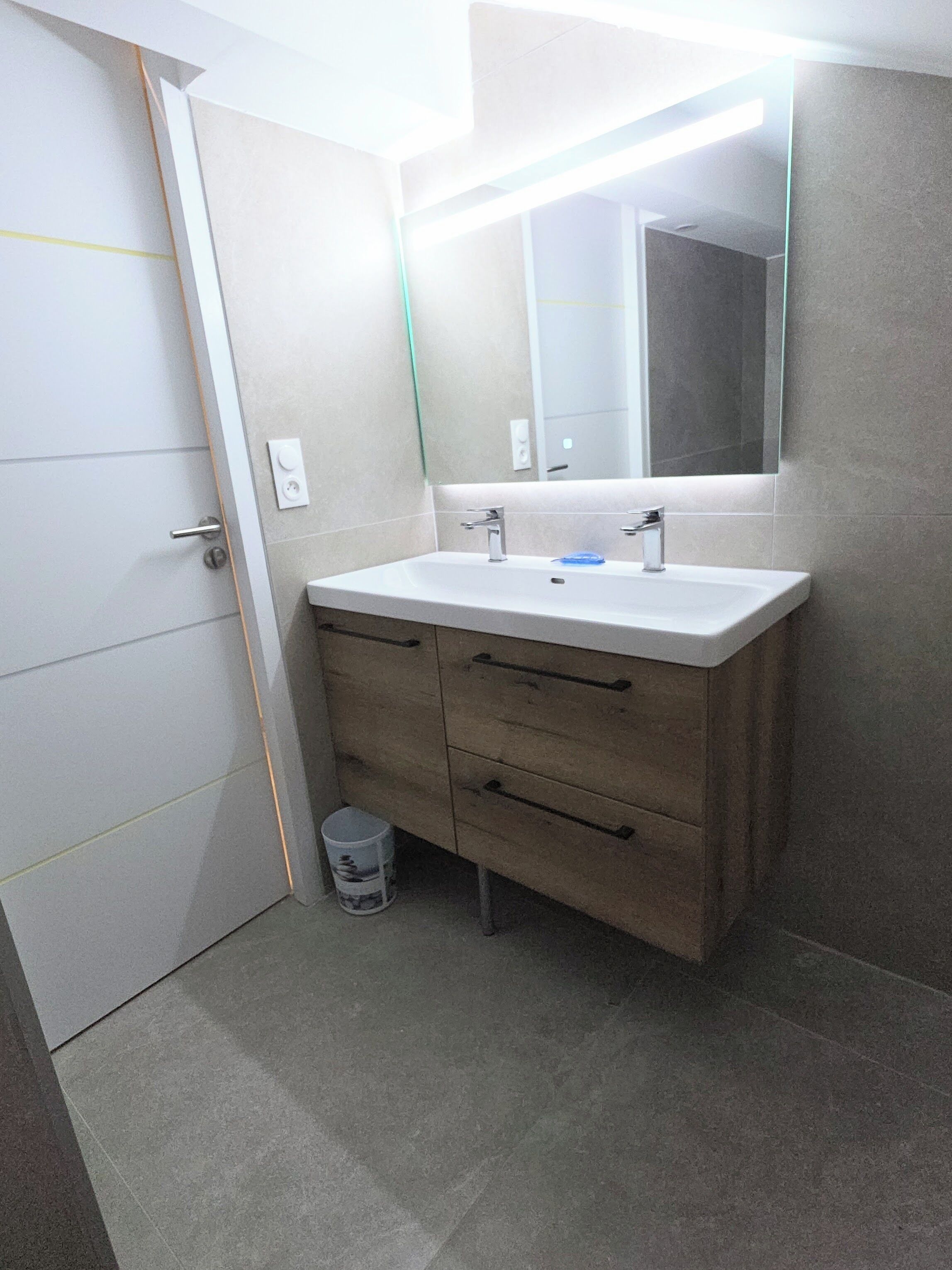 Une salle de bain avec un lavabo blanc, deux tiroirs, un miroir et une poubelle.