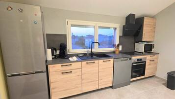 Eine moderne Küche mit einem Edelstahlkühlschrank, Holzschränken, einem schwarzen Geschirrspüler, einer Mikrowelle und einem Fenster mit Blick auf Häuser.