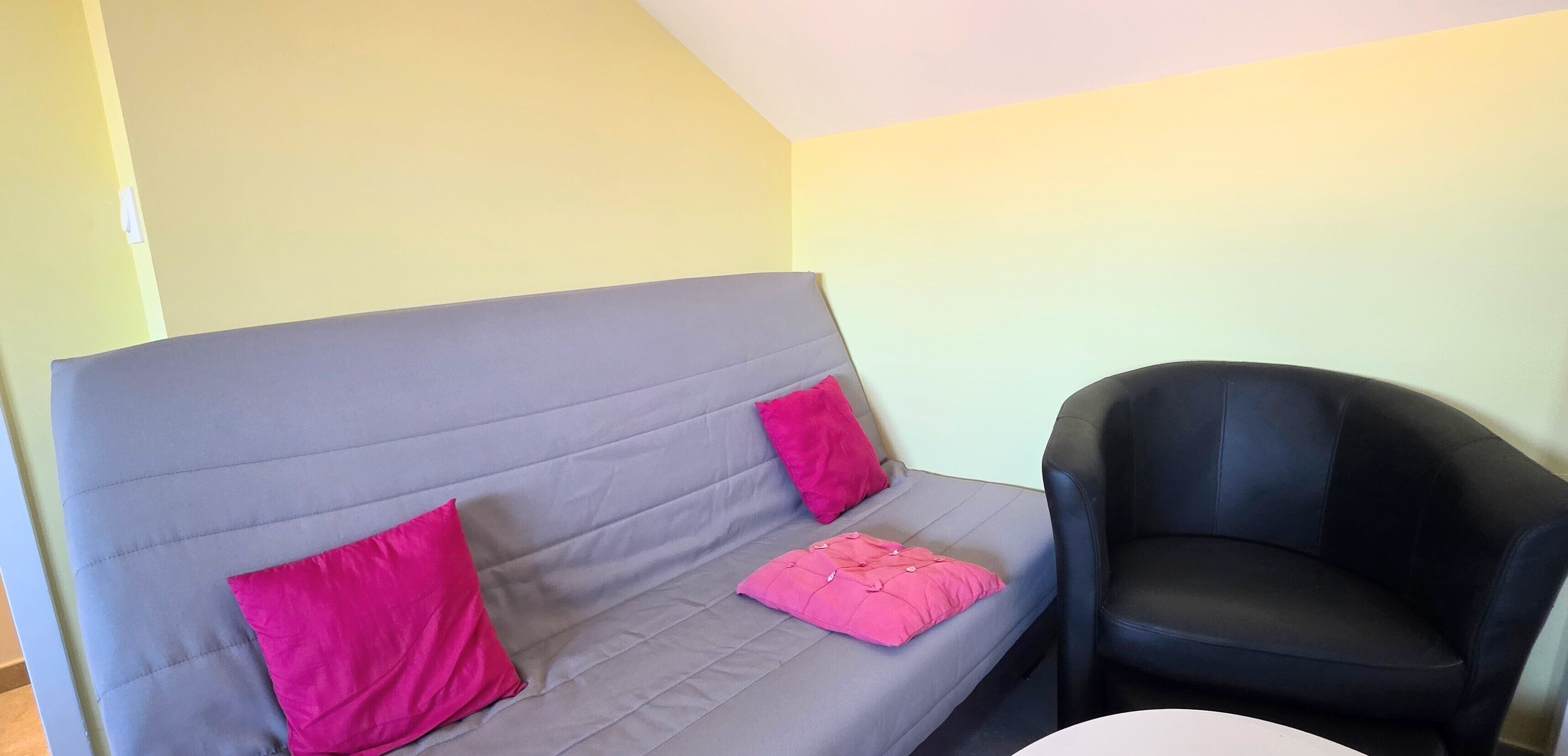 A grey sofa with pink cushions, a black armchair, and a white table in a room with light yellow walls.