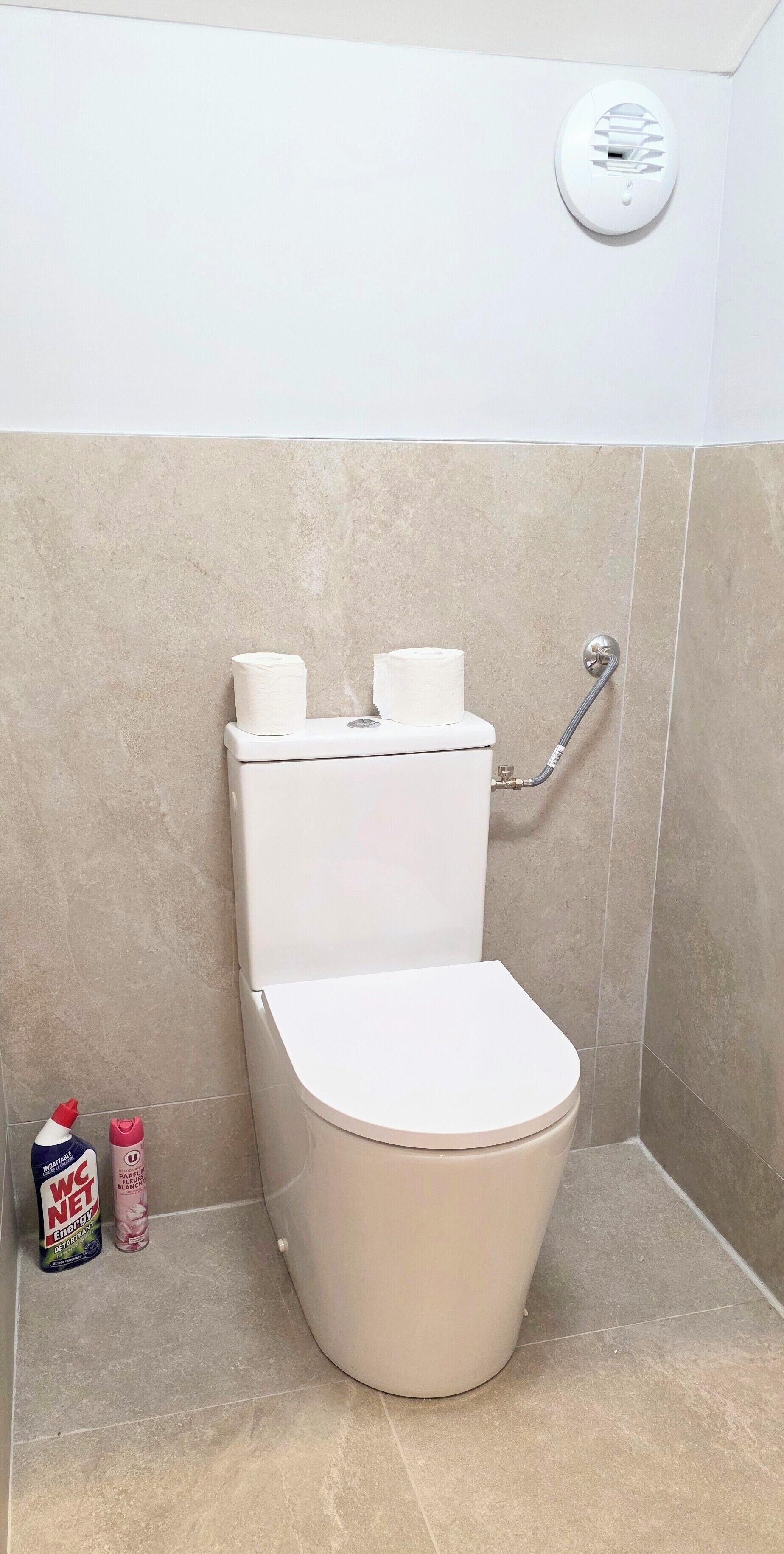 A bathroom with a toilet, two rolls of toilet paper, a shower hose, and two toiletry bottles.