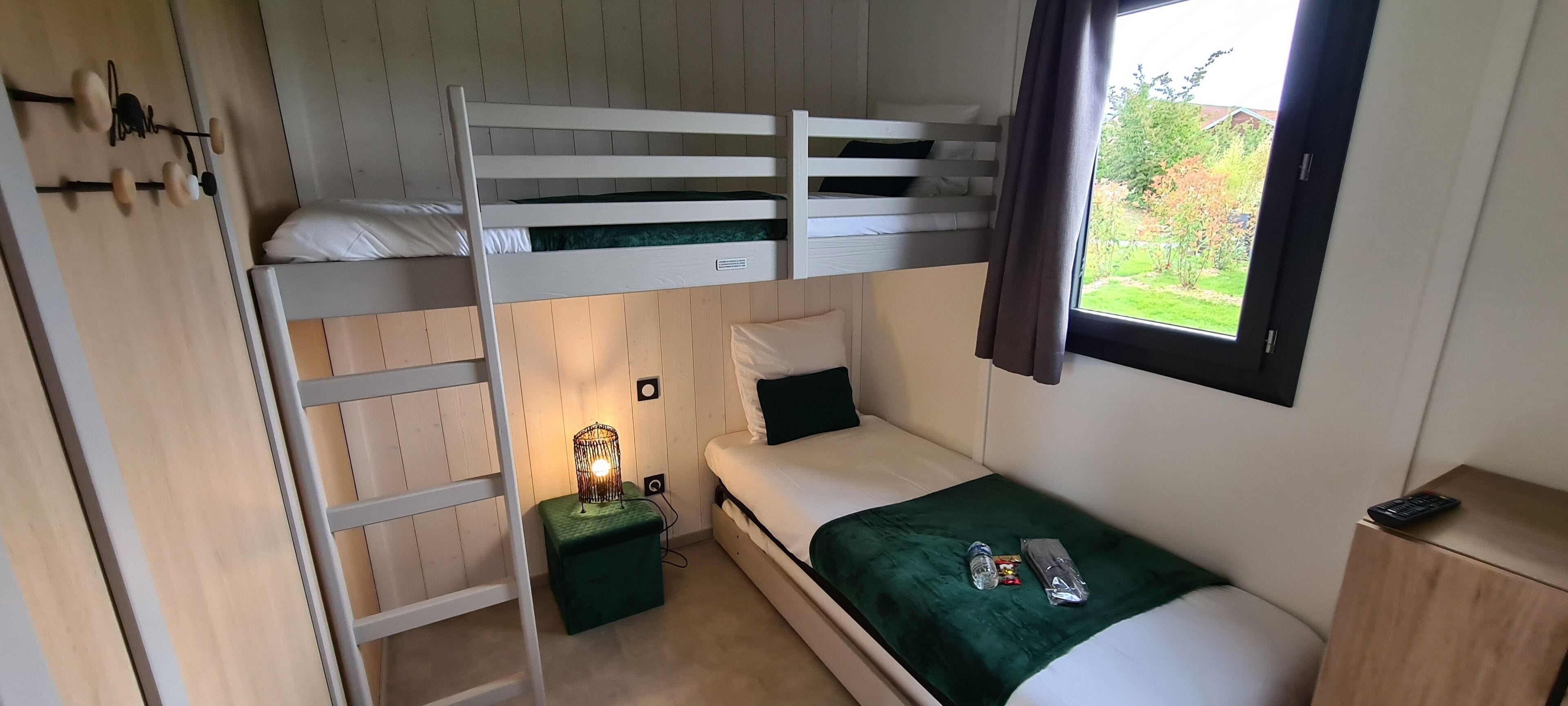 A bunk bed room with a green cushion, a bedside table with a lamp, and a window with a view of greenery.
