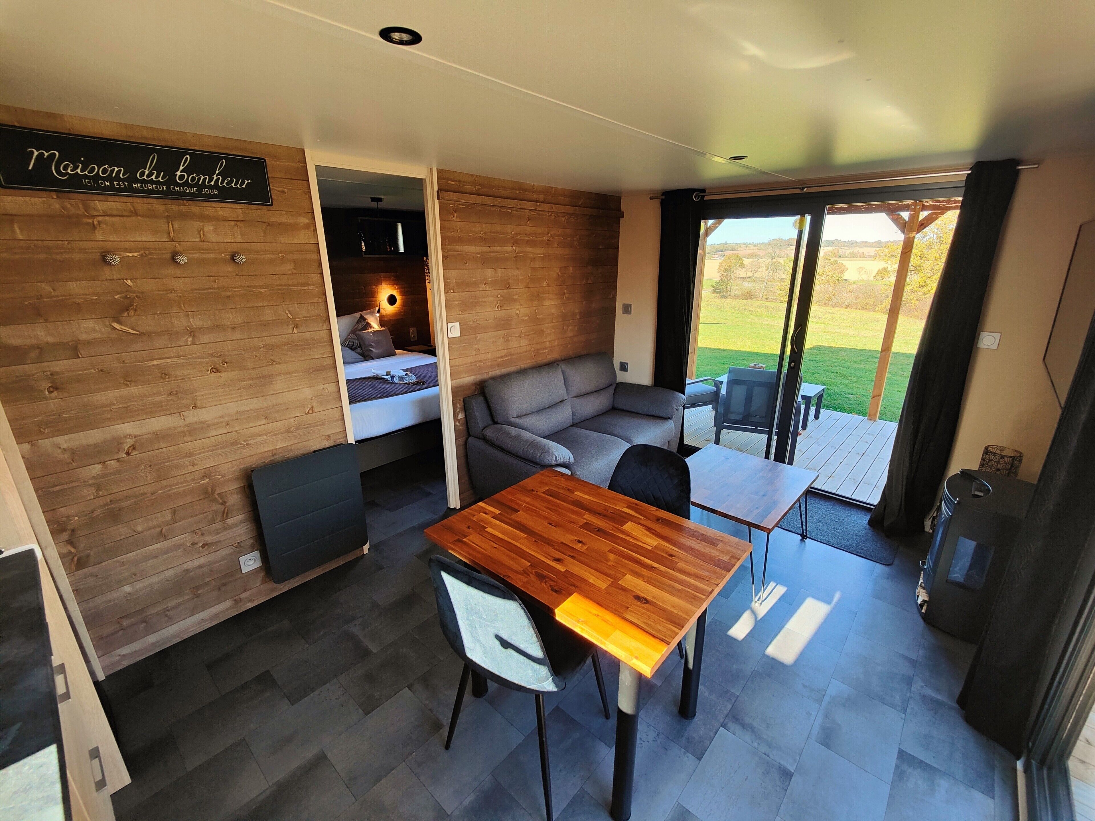 A compact, modern room with a wooden wall, a bed, a gray sofa, a wooden table, and a dining area.