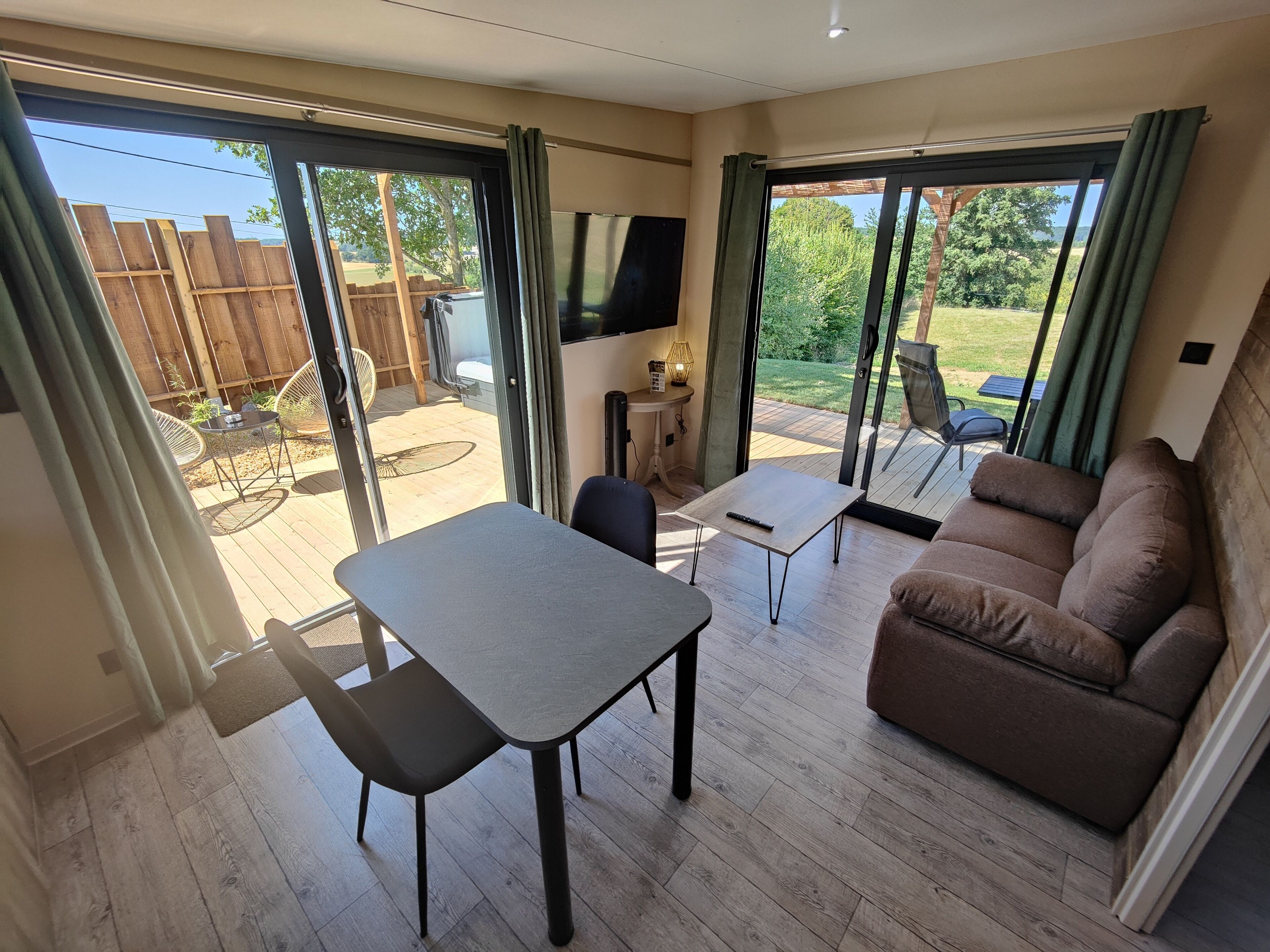 Un vaste espace de vie comprenant un canapé, une petite table et un fauteuil. Une porte-fenêtre coulissante donne accès à un patio extérieur doté d’une terrasse en bois et d’un téléviseur fixé au mur.