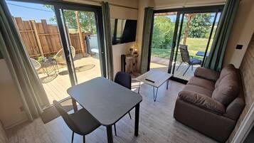 A spacious living area with a sofa, a small table, and a chair. There is a sliding glass door leading to an outdoor patio with a wooden deck and a TV mounted on the wall.