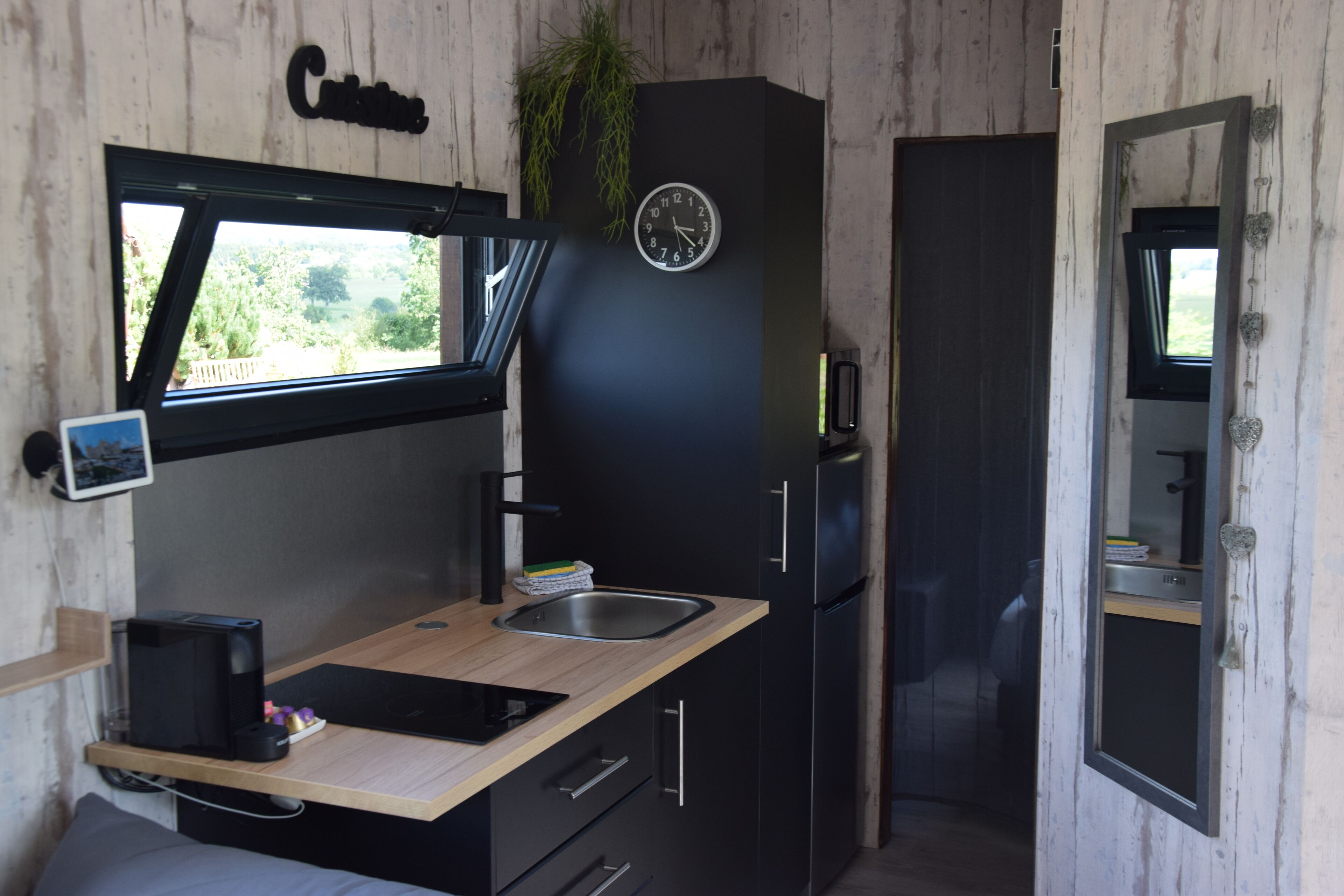 A compact kitchen with a built-in sink, a clock, and a window overlooking greenery.