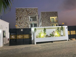 Un bâtiment moderne doté d’une façade en pierre, d’une entrée sécurisée et d’un espace paysager agrémenté de plantes et d’un élément décoratif aquatique.