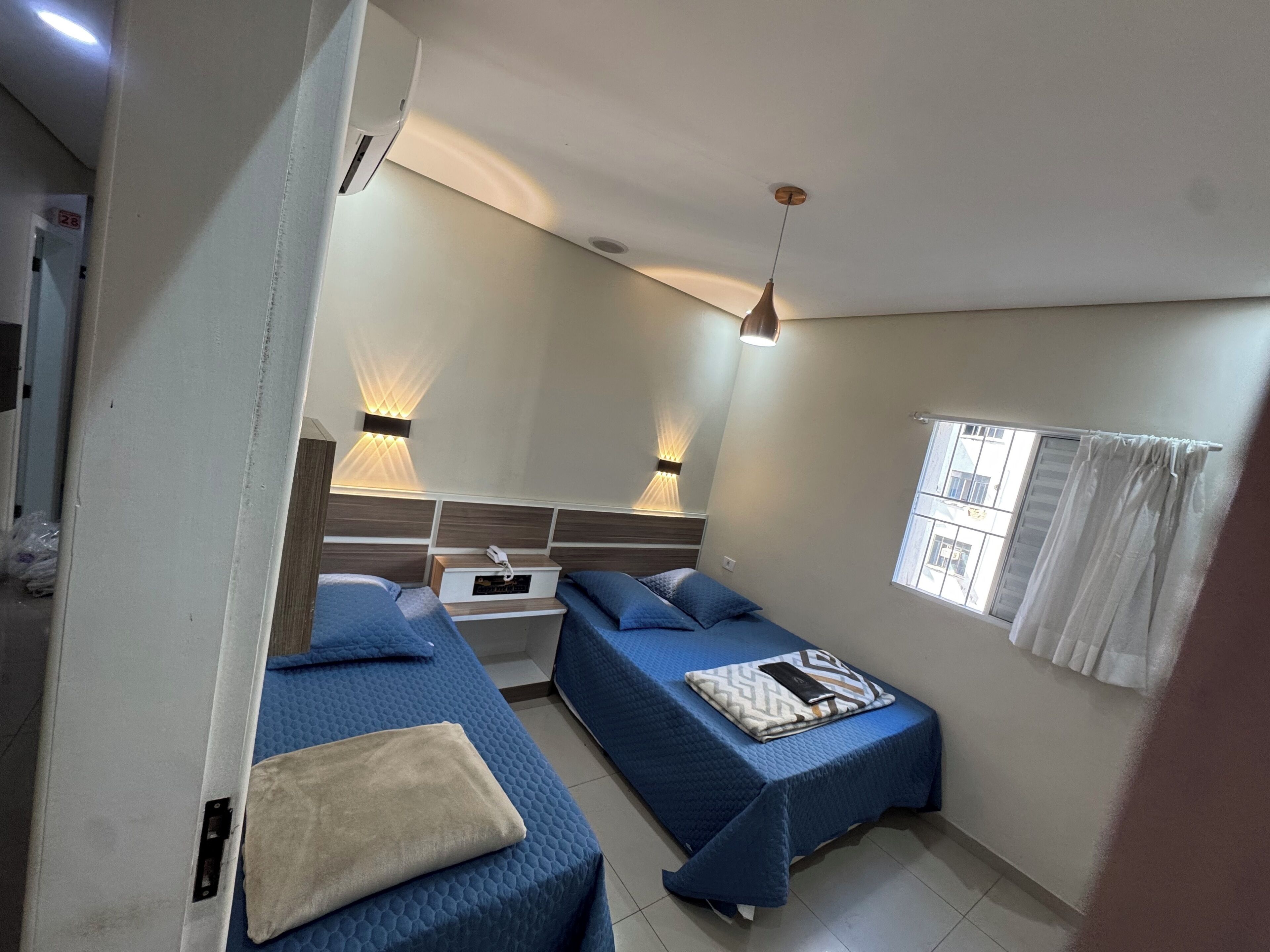 Una habitación con dos camas, una mesita de noche, una lámpara de pared, una ventana con cortinas y una lámpara en el techo.