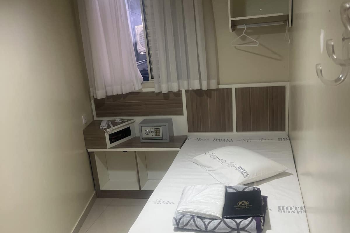 A hotel room with a bed, a bedside table with a phone and radio, a window with curtains, and a wall-mounted shelf.