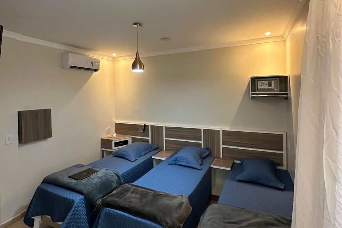 A room with four beds, a wall-mounted air conditioner, a bedside table with a lamp, and a ceiling light fixture.