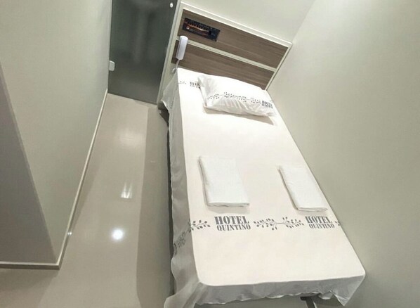 A single bed with a white bedspread and two pillows, a bedside table with a phone, and a small shelf with a book. - Hotel Quintino (São Paulo)