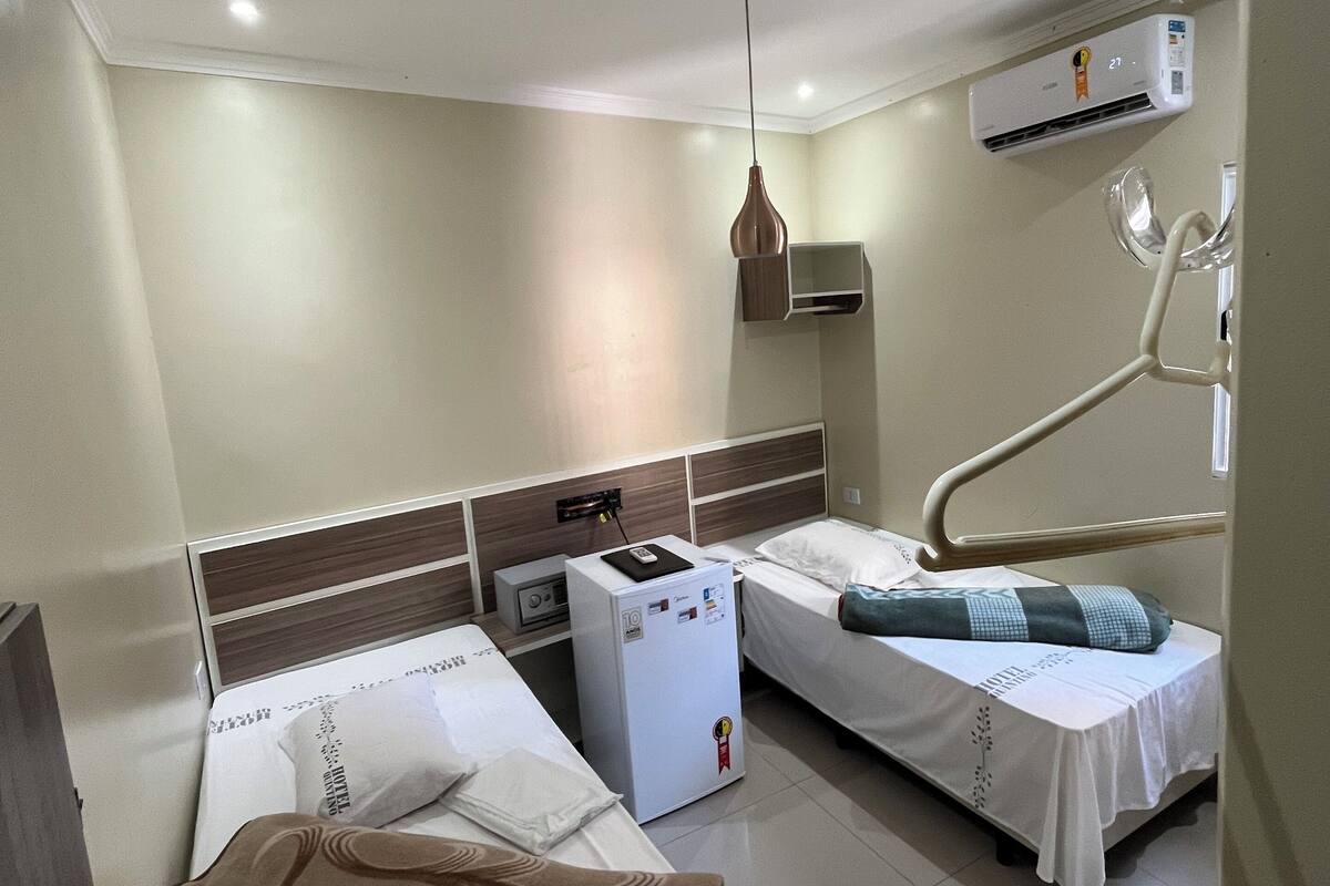 A hotel room with two single beds, a small bedside table, a wall-mounted air conditioner, and a ceiling light fixture.