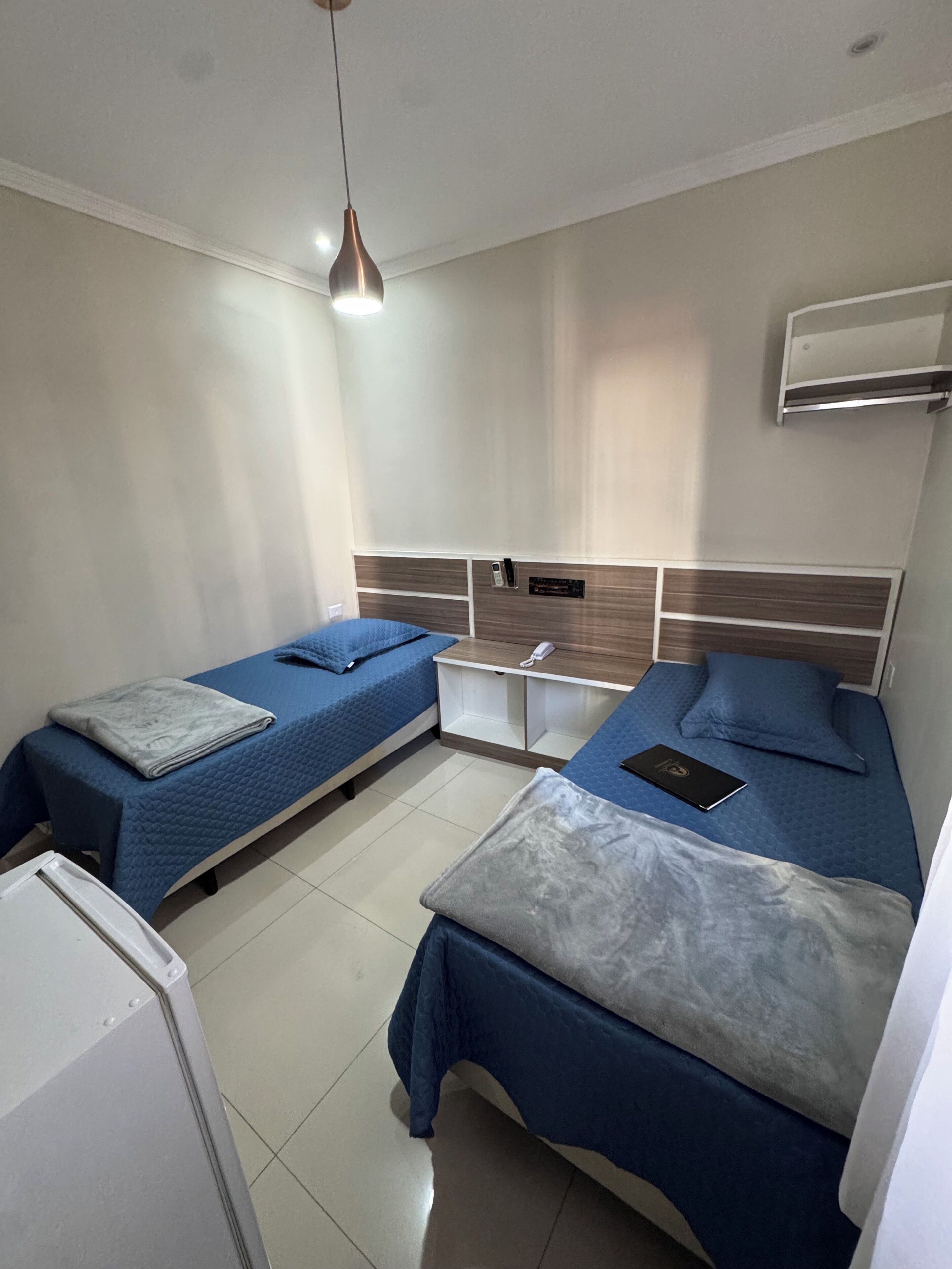A hotel room with two beds, a small table, a wall-mounted air conditioner, and a ceiling light.