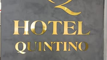 Ein Schild mit der Aufschrift 'HQ HOTEL QUINTINO' in goldener Schrift auf grauem Grund.