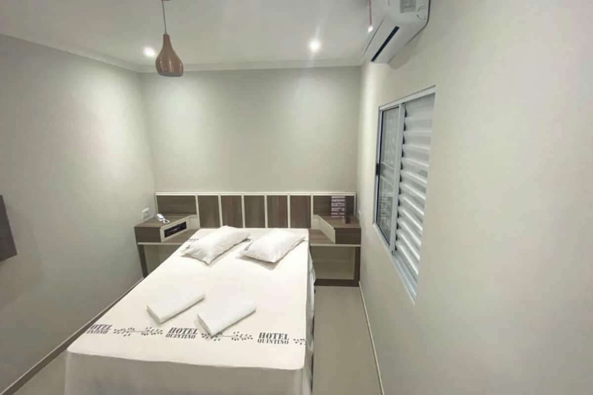 A hotel room with a bed, bedside table, and a window with blinds.