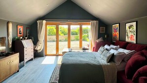 A bedroom with a bed, a red sofa, a wooden cabinet, and a sliding glass door leading to a garden. - Eclectic Barn · Pergola & Dog-Friendly Garden (Calne)