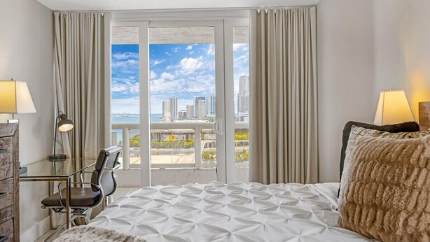 A hotel room with a bed, a desk, a chair, and a view of a cityscape through large windows. - Bay Overlooking Condo | Downtown | StayCozy (Miami)