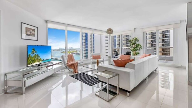 A modern living room with a large TV, white furniture, and a view of the city. - Bay View High-Rise | Private Balcony | StayCozy (Miami)