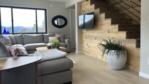 A modern living room with a grey sofa, a coffee table, and a large potted plant. A TV is mounted on a wooden wall above a staircase. -  Off Grid Mountain Retreat w/ Amazing Views (Idaho Springs)