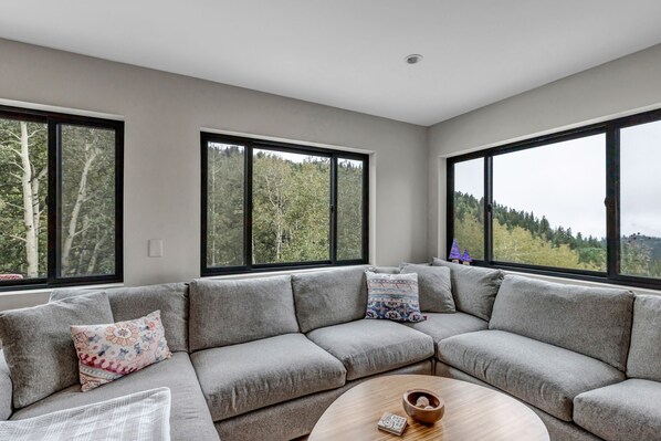 A modern living room with a grey sectional sofa, a round wooden coffee table, and large windows offering a view of a forested area. - Off Grid Mountain Retreat w/ Amazing Views (Idaho Springs)