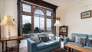 Ein Wohnzimmer mit einem blauen Sofa, einem Holztisch und einem gemusterten Teppich. Ein Bücherregal und ein Fenster mit Blick auf die Stadt sind ebenfalls vorhanden.