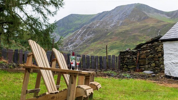 A wooden chair with a bottle and glass on it, set in a grassy area with a stone wall and a white building in the background. - Ceunant Bach - One Bedroom Cottage, Sleeps 2 (Bethesda)